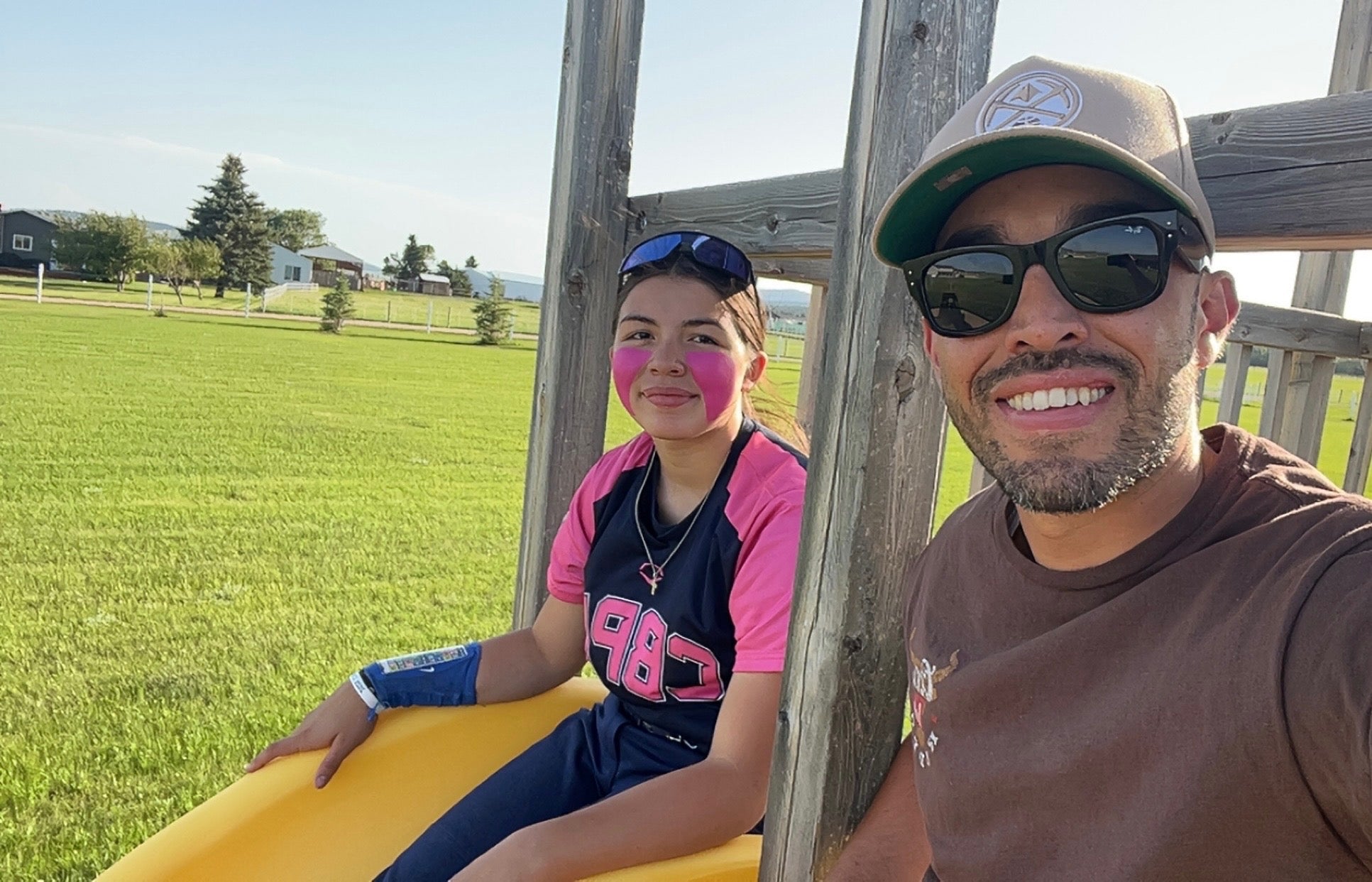 Man and child sitting on a yellow slide in an outdoor setting with grass and trees in the background.
