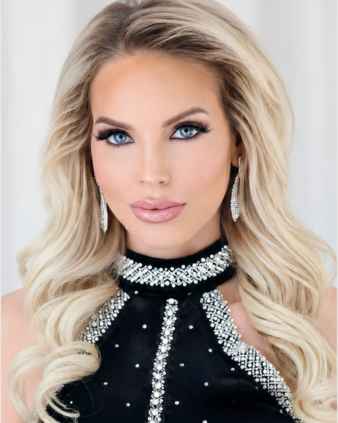 A close-up portrait of a blonde woman with long wavy hair, wearing a black embellished halter-neck top and matching dangling earrings against a soft light background.