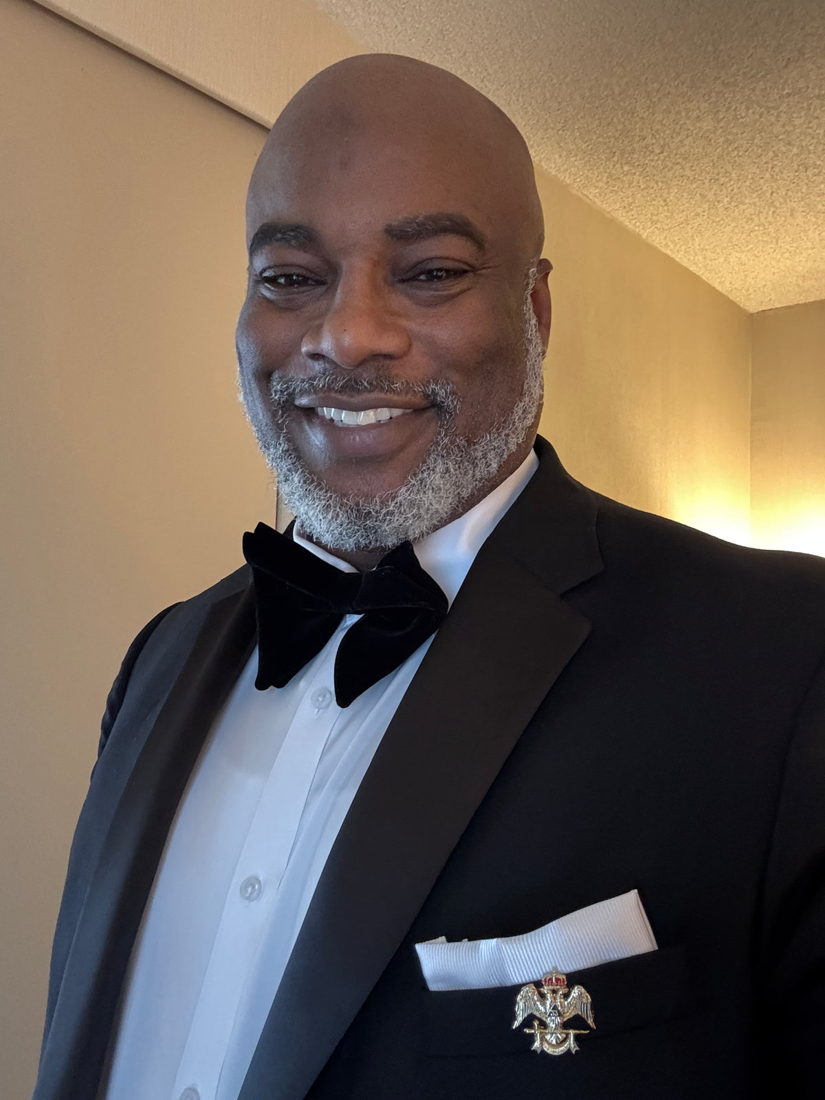 A smiling bald man with a gray beard wearing a black tuxedo jacket, white dress shirt, and black bow tie, with a white pocket square and a small lapel pin, photographed indoors against a beige wall.