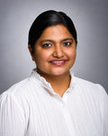 A professional portrait of a woman with dark hair, wearing a white pleated button-up shirt, set against a solid gray studio background.