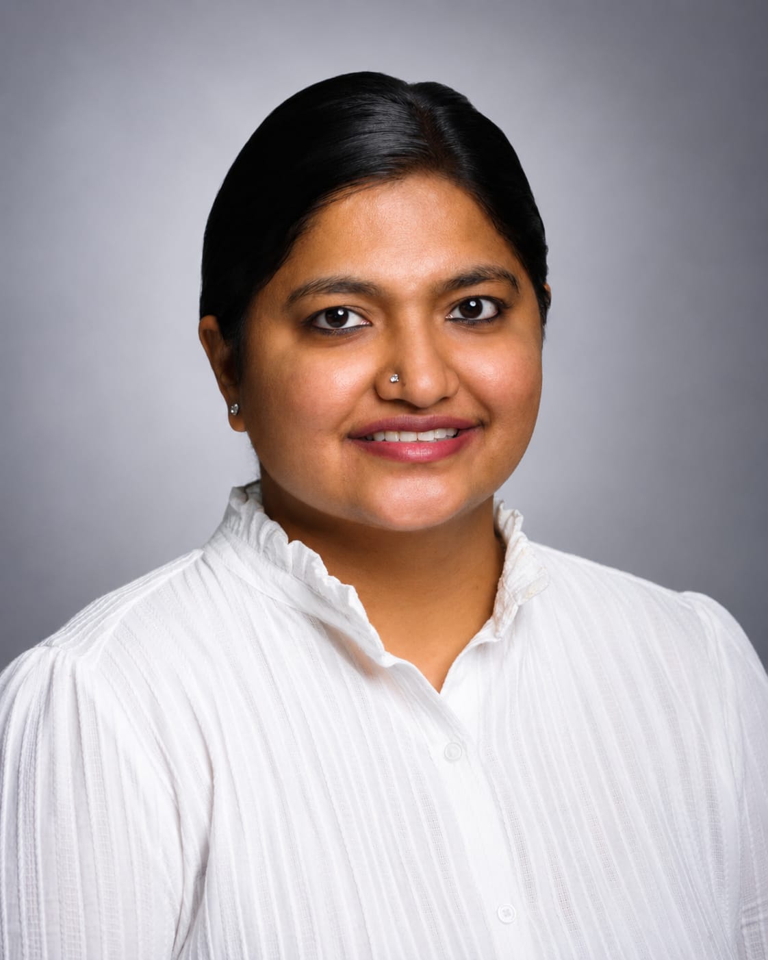 A professional portrait of a woman with dark hair, wearing a white pleated button-up shirt, set against a solid gray studio background.