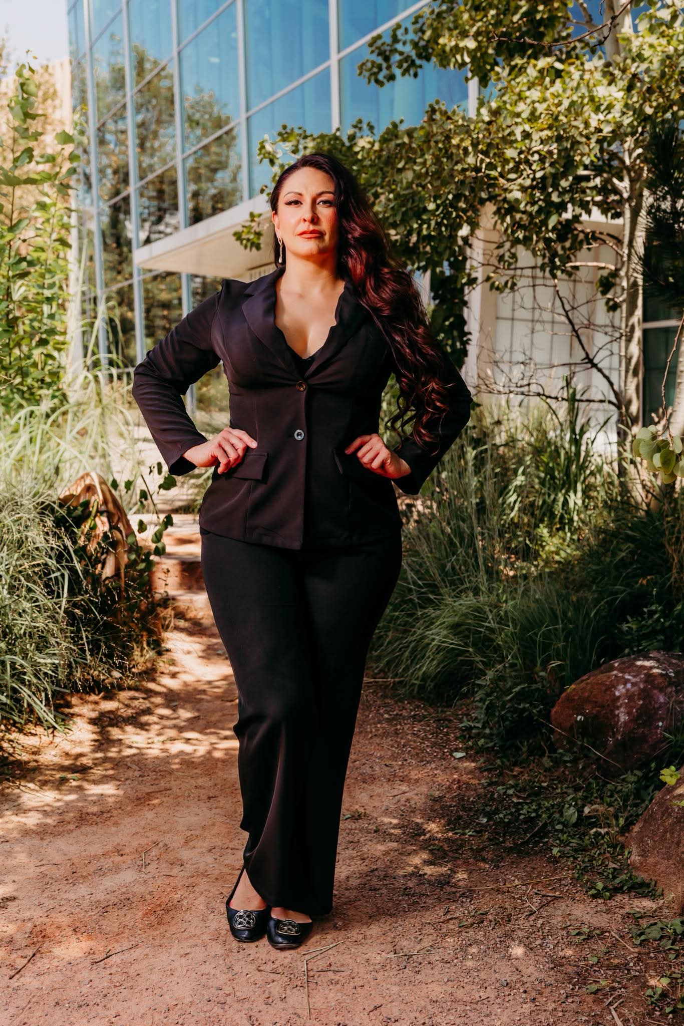 Woman in a black suit standing outdoors with greenery and a building in the background