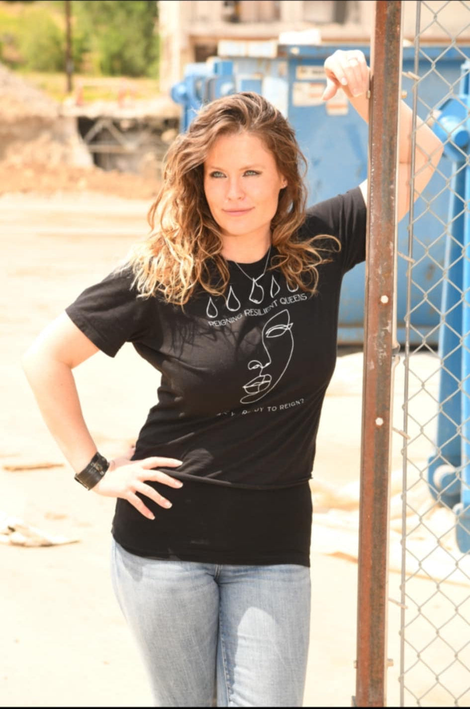 Woman wearing a black t-shirt with text, standing outdoors near a chain-link fence.