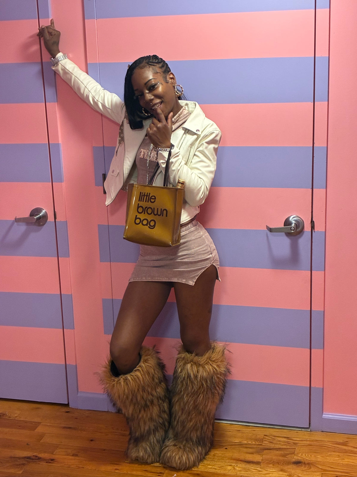 A woman posing in front of pink and purple striped doors, wearing a white jacket, a light pink mini skirt, and oversized brown faux fur boots, while holding a gold 'little brown bag' tote.