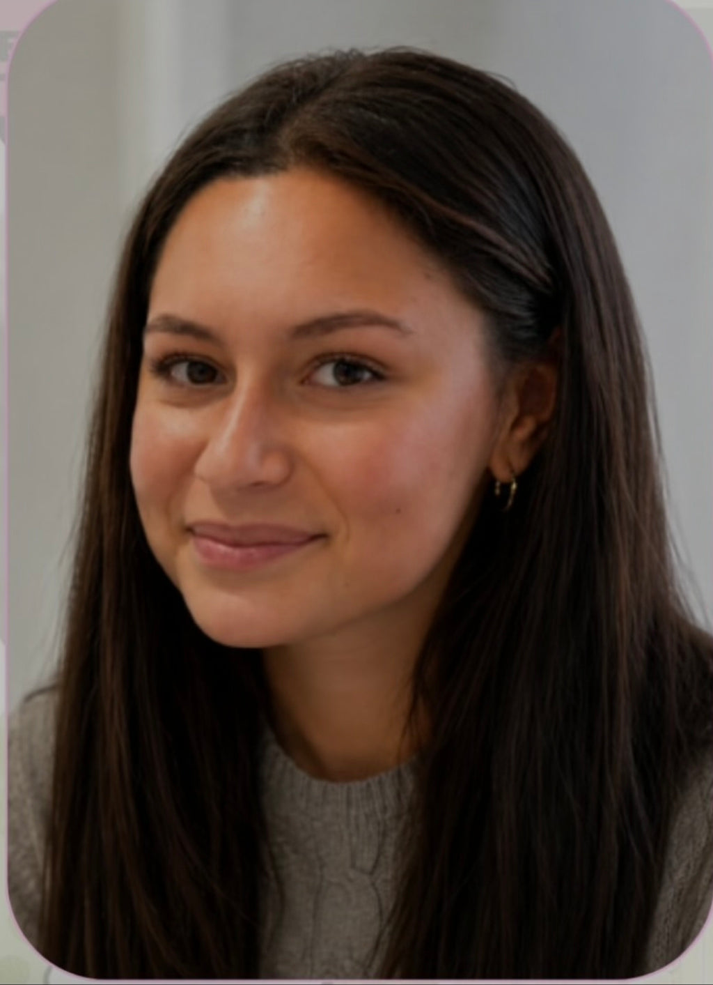A close-up portrait of a woman with long dark brown hair, wearing a gray knit top and a small gold hoop earring, set against a soft neutral background with rounded corners.