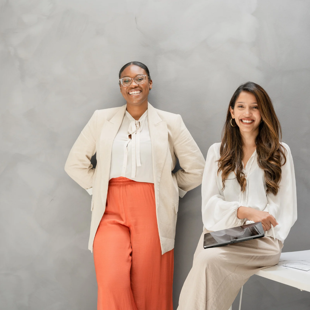 Two women standing against a gray background, one holding a tablet.