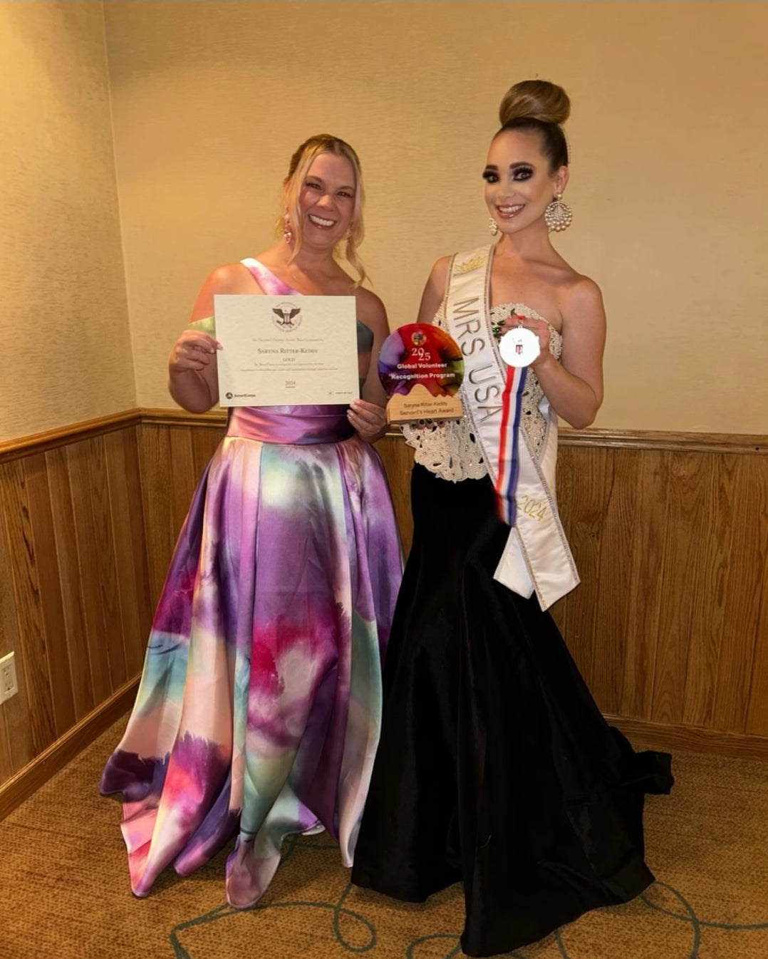 Two women in formal attire, one holding a certificate and the other with a sash, in an indoor setting.