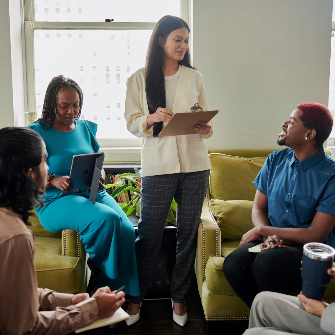 Group therapy session with a facilitator taking notes in a bright room.