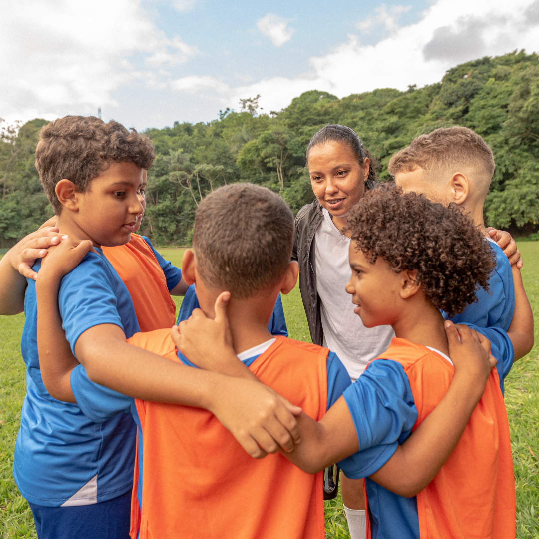 Group of children in sports jerseys huddled together on a grassy field with trees in the background